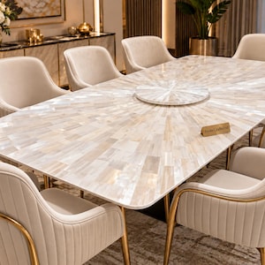 May include: A rectangular dining table featuring a white mother-of-pearl inlay surface with a sunburst pattern, a matching circular lazy Susan, and a small wooden sign labelled StoneCraftsInd, surrounded by eight cream-coloured upholstered chairs.