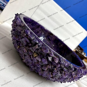 May include: A handcrafted amethyst bowl. The exterior is encrusted with raw amethyst crystals, while the interior has a polished, smooth surface. The bowl's deep purple color is accentuated by the natural variations in the stone.