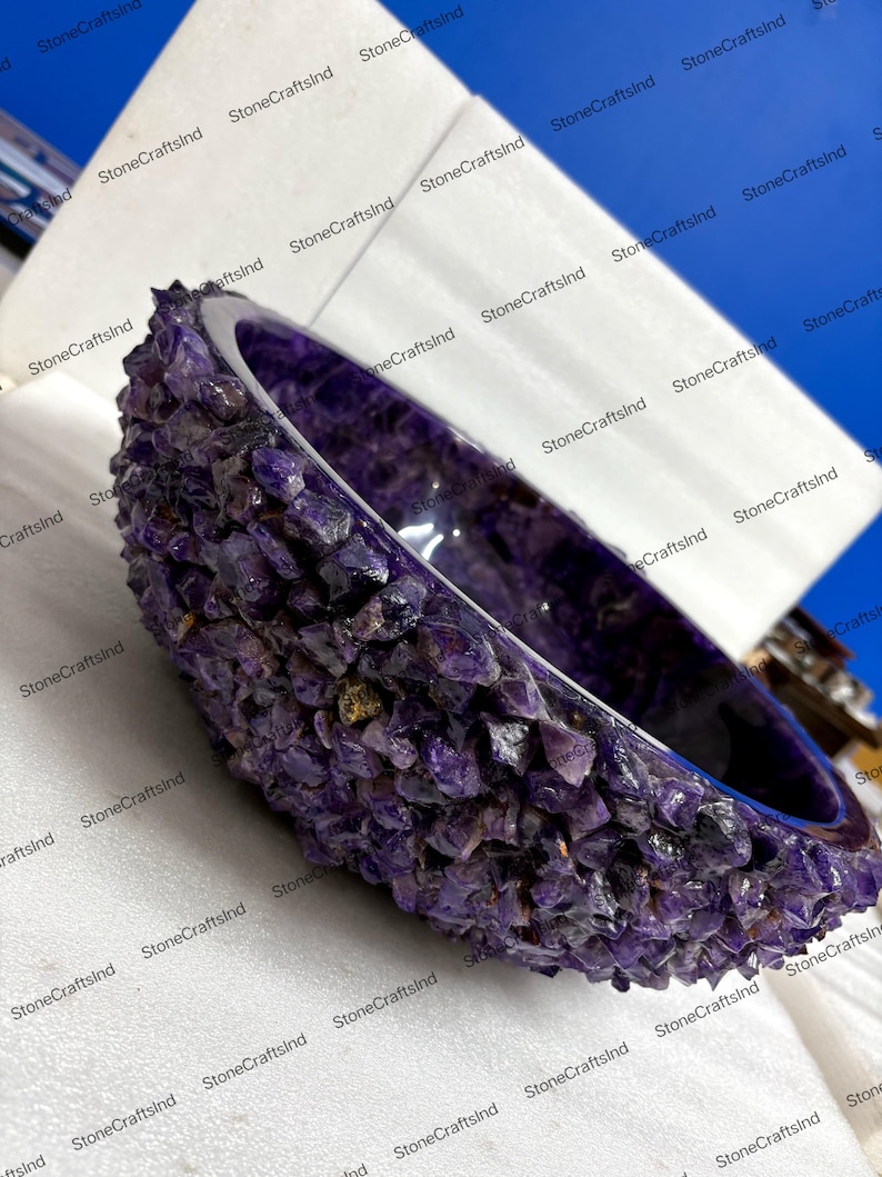May include: A handcrafted amethyst bowl. The exterior is encrusted with raw amethyst crystals, while the interior has a polished, smooth surface. The bowl's deep purple color is accentuated by the natural variations in the stone.