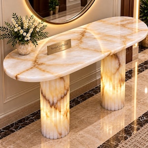 Op de afbeelding: Een ovale consoletafel gemaakt van doorschijnende witte en amberkleurige onyxsteen met een gepolijste afwerking. De tafel heeft twee gecanneleerde cilindrische zuilvoeten en een glad bovenblad. Een klein gouden plaatje vermeldt StoneCraftsInd.