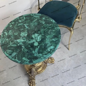 Op de afbeelding: Een ronde groene malachiet tafelblad met gouden metalen poten en een gouden metalen basis. Het tafelblad is gemaakt van veel kleine stukjes malachiet die in een moza&iuml;ekpatroon zijn gerangschikt. De tafel staat op een lichtgrijze vloer.