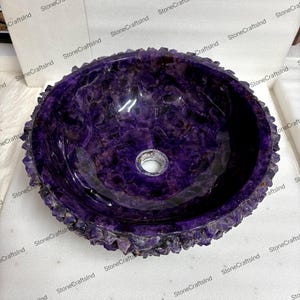 May include: A round, purple amethyst stone sink with a rough, textured edge. The interior is smooth and polished, featuring a drain hole in the center. The sink is a vibrant shade of purple, showcasing the natural beauty of the stone.
