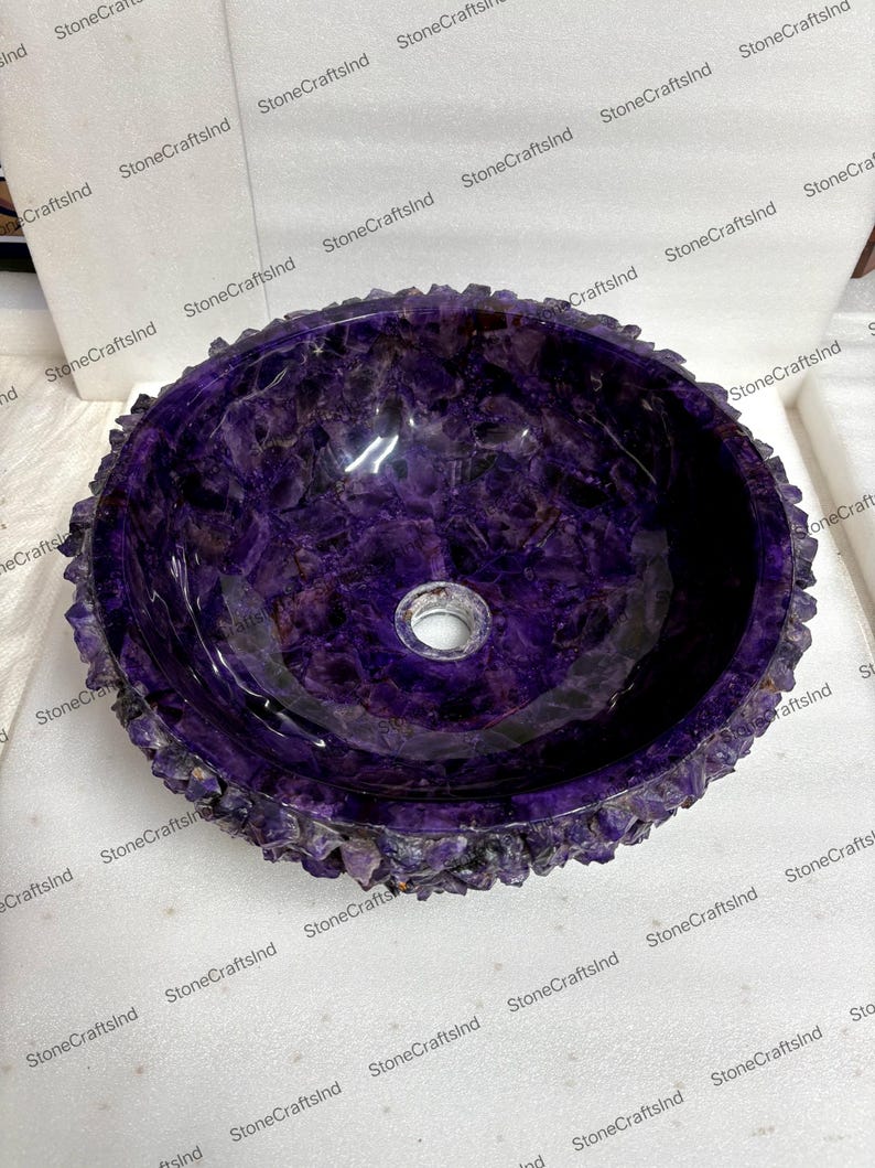 May include: A round, purple amethyst stone sink with a rough, textured edge. The interior is smooth and polished, featuring a drain hole in the center. The sink is a vibrant shade of purple, showcasing the natural beauty of the stone.