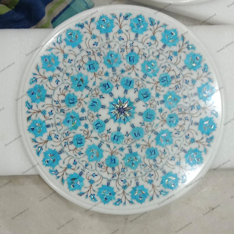 May include: A round, white marble tabletop with an intricate floral design. The design features turquoise and silver-colored inlays arranged in a symmetrical pattern. The flowers are surrounded by delicate, dark blue vines and leaves. The tabletop has a smooth, polished finish.