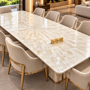 Op de afbeelding: Een rechthoekige eettafel met een gepolijst oppervlak van ingelegde crèmekleurige stenen tegels in een zonnestraalpatroon, ondersteund door een bijpassende stenen voet, omringd door acht beige gestoffeerde stoelen met gouden metalen frames.