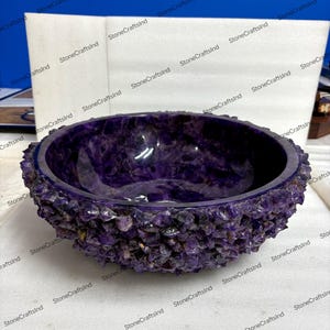May include: A handcrafted amethyst bowl with a rough, textured exterior and a smooth, polished interior. The bowl is a deep purple color, showcasing the natural beauty of the gemstone. This decorative bowl is ideal for home decor.