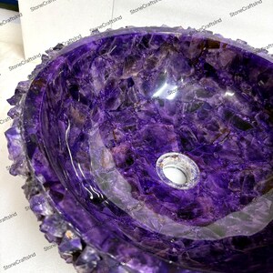 May include: A polished, oval-shaped amethyst stone sink. The sink's interior is a deep purple with crystalline patterns, and the rim features a rough, textured edge. A drain hole is visible in the center.