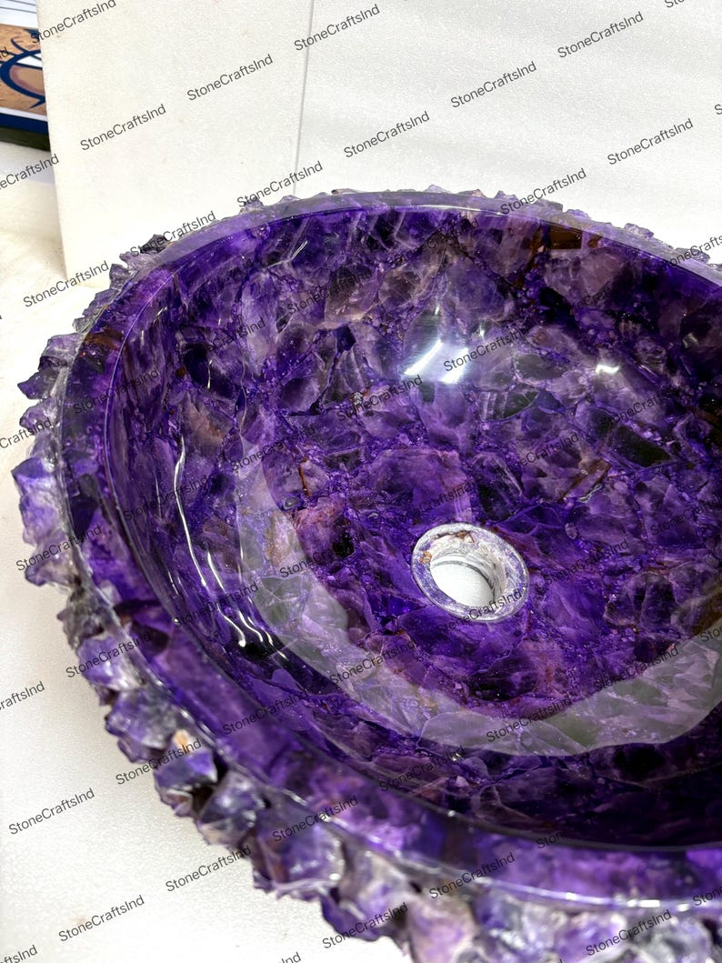 May include: A polished, oval-shaped amethyst stone sink. The sink's interior is a deep purple with crystalline patterns, and the rim features a rough, textured edge. A drain hole is visible in the center.