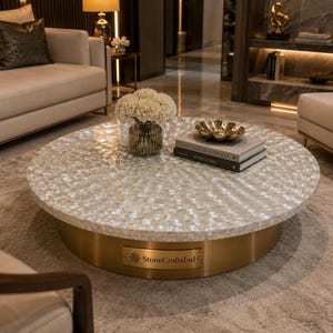 May include: A round coffee table featuring a white mother-of-pearl tiled top and a cylindrical brass base. A rectangular metal plaque on the base displays the text StoneCraftsInd. The table surface holds a glass vase with white flowers.