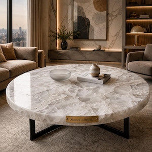 May include: A round coffee table featuring a polished white quartz stone top with natural crystalline textures and a black metal base. A small brass plaque on the edge reads StoneCraftsInd. A glass bowl and a book titled Natures Gemstones sit on top.