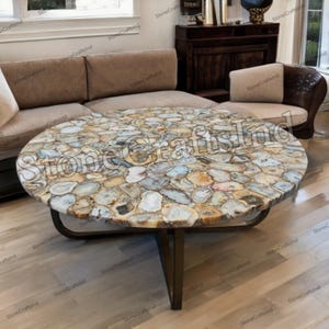 May include: A round coffee table with a mosaic top made of various colored stones. The stones are arranged in a pattern, with shades of blue, beige, and gray. The table has a dark metal base.