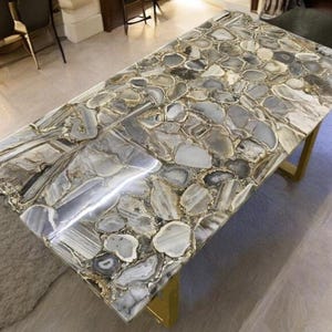 May include: A rectangular table with a mosaic-style top made of gray, white, and brown stones. The table has a gold-colored metal base. The stone top is polished and reflects light, creating a luxurious look. The table is a statement piece.