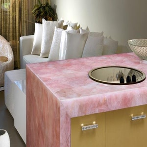 May include: A pink quartz worktop with a built-in hob and a mirrored surface. The worktop is part of a kitchen island with gold cabinets and crystal drawer pulls.