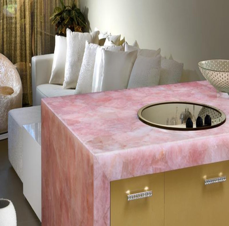 May include: A pink quartz worktop with a built-in hob and a mirrored surface. The worktop is part of a kitchen island with gold cabinets and crystal drawer pulls.