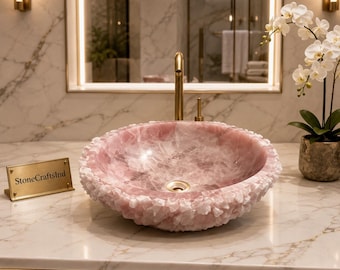 May include: A round vessel sink crafted from polished pink rose quartz with a rough, crystalline exterior texture. The sink features a central drain and is accompanied by a small gold-toned plaque engraved with the text StoneCraftsInd.