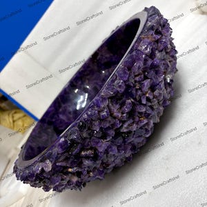 May include: A handcrafted, amethyst stone bowl. The bowl is a deep purple color, with a rough, textured exterior made of amethyst crystals. The interior is smooth and polished. The bowl is a unique decorative item.
