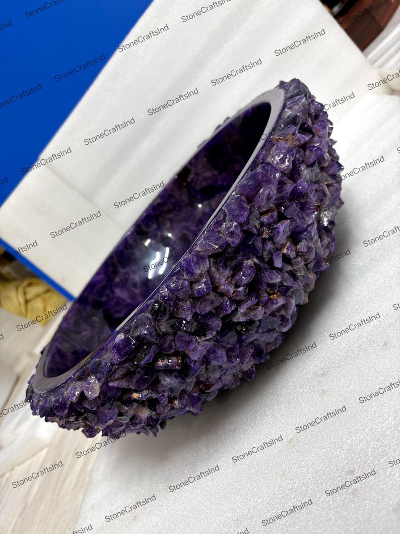 May include: A handcrafted, amethyst stone bowl. The bowl is a deep purple color, with a rough, textured exterior made of amethyst crystals. The interior is smooth and polished. The bowl is a unique decorative item.