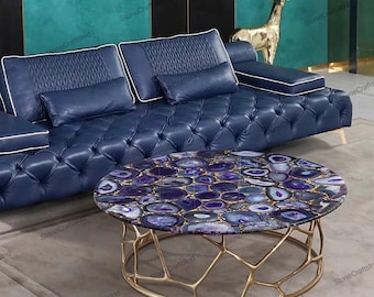 May include: A round coffee table with a gold metal frame and a purple agate stone top. The table is in front of a blue leather sofa with white piping.