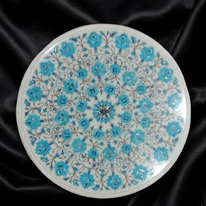 May include: Round white marble table top with a detailed floral inlay design. The design features turquoise and blue floral patterns radiating from a central starburst. The table top is set against a black background.