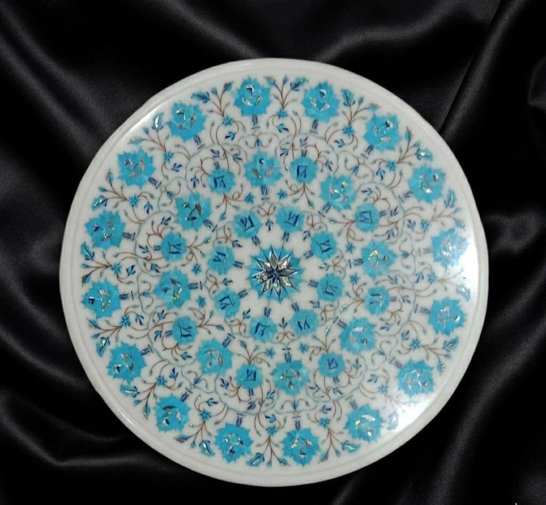 May include: Round white marble table top with a detailed floral inlay design. The design features turquoise and blue floral patterns radiating from a central starburst. The table top is set against a black background.