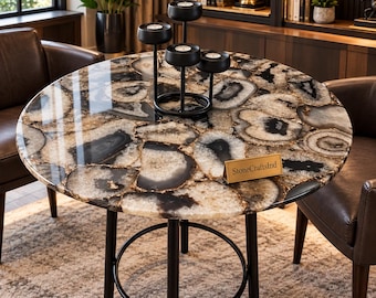 May include: A round table featuring a polished surface made of inlaid black and beige agate slices. The table has four black metal legs connected by a circular support ring. A black metal candle holder and a gold sign reading StoneCraftsInd sit on top.