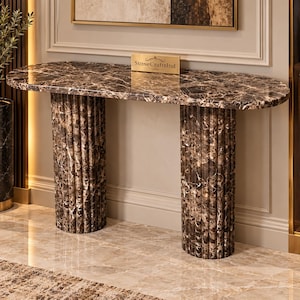 Op de afbeelding: Een console-tafel van bruin en wit marmer met een ovaal blad en twee gecanneleerde cilindrische poten. Een klein rechthoekig goudkleurig plaatje met de tekst StoneCraftsInd staat in het midden van het tafelblad, met een gepolijste afwerking.