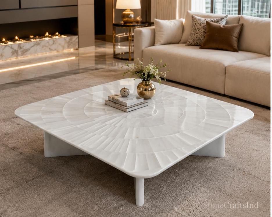 Op de afbeelding: Een vierkante witte salontafel met een uniek, getextureerd oppervlakteontwerp. De tafel heeft afgeronde hoeken en staat op een stevige basis met vier poten. Decoratieve items, waaronder boeken en een vaas, zijn op de tafel geplaatst. Het is een blikvanger in een moderne woonkamer.