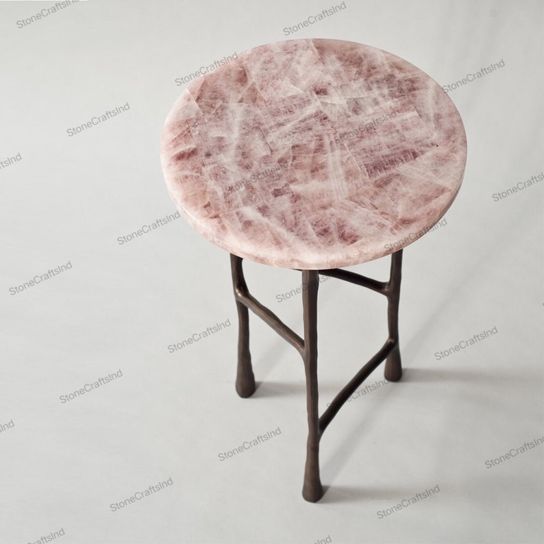 May include: A round pink quartz table top with a metal base. The base has four legs that are curved and have a rustic finish.