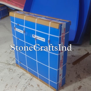 May include: A blue cardboard box with a brown top is wrapped in white tape and labelled "FRAGILE HANDLE WITH CARE". The box is sitting on a concrete floor. The text "StoneCraftsInd" is visible on the box.