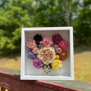 Puede incluir: Una caja de sombra enmarcada en blanco con una variedad de flores prensadas en tonos de rosa, morado, amarillo y naranja. Las flores están dispuestas en forma de corazón.