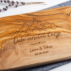 Könnte beinhalten: Holzschneidebrett mit eingraviertem Bergketten-Design und dem Schriftzug "Liebe versetzt Berge". Darunter die Namen "Laura & Tobias" und das Datum "03.08.2024". Das Brett hat eine natürliche Holzmaserung.