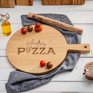 Könnte beinhalten: Ein rundes, hölzernes Pizzabrett mit Griff. Das Brett ist mit dem Text "Andris' PIZZA" und einer kleinen Pizzastückgrafik graviert. Das Brett liegt auf einem grauen Tuch mit roten Kirschtomaten.