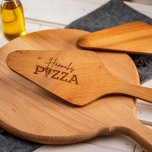 Könnte beinhalten: Ein Holzpizza-Schaufel mit einem Griff und dem Text "Hannah's PIZZA" auf der Oberfläche graviert. Die Schaufel liegt auf einem Holzpizza-Servierbrett.