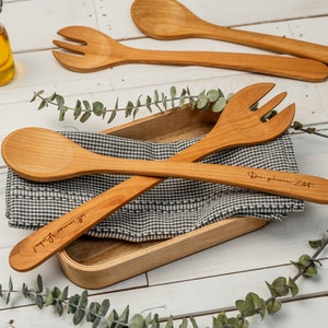 Op de afbeelding: Twee houten keukengerei, een lepel en een vork, zijn gekruist en geplaatst in een houten doos bekleed met een zwart-wit geblokt doek. De keukengerei zijn gegraveerd met de tekst "Die gemeinsame Zeit..." en "...ist immer schön!"
