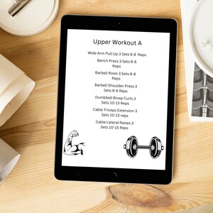 May include: A digital tablet displaying a workout routine titled "Upper Workout A". The routine includes exercises such as wide arm pull ups, bench press, barbell rows, barbell shoulder press, dumbbell bicep curls, cable triceps extension, and cable lateral raises. The number of sets and reps for each exercise is listed.