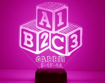May include: A pink acrylic night light shaped like a stack of blocks with the letters A1, B2, and C3. The base of the night light is black and has the name GABBIE and the date 5-17-16.