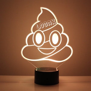May include: A white outline of a smiling poop emoji with eyes and a name "LOGAN" on top. The emoji is a night light with a black base.