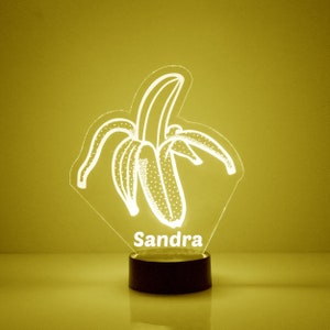May include: A white illuminated acrylic night light shaped like a banana with the name "Sandra" printed below. The night light is on a black base.