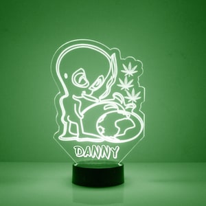 May include: A white LED night light with a cartoon alien holding a globe and marijuana leaves. The light has a black base and the name "DANNY" is printed on the front.