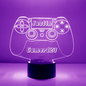 May include: A glowing, purple-hued LED lamp shaped like a video game controller. The controller design features the name "Justin" and the text "Gamer123". The lamp sits on a black base, creating a striking visual effect.