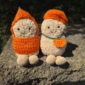 Könnte beinhalten: Zwei Plüschpuppen mit beigen, strukturierten Körpern und kleinen runden Füßen. Die linke Puppe trägt eine orangefarbene Häkelkapuze und einen passenden Pullover. Die rechte Puppe trägt ein orangefarbenes Häkelbére und eine grüne Kette mit Anhänger.