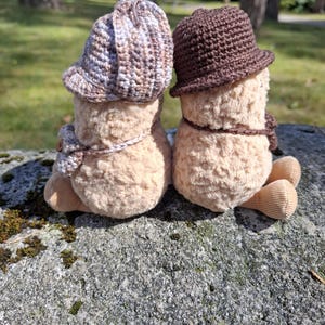 Puede incluir: Dos figuras de peluche con sombreros y bufandas de ganchillo sentadas en una roca. Una figura lleva un sombrero y una bufanda marrones y beiges, la otra un sombrero y una bufanda marrones. Las figuras son de color beige claro con pies de pana.