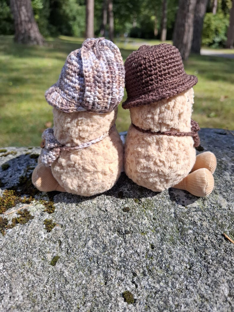 Puede incluir: Dos figuras de peluche con sombreros y bufandas de ganchillo sentadas en una roca. Una figura lleva un sombrero y una bufanda marrones y beiges, la otra un sombrero y una bufanda marrones. Las figuras son de color beige claro con pies de pana.
