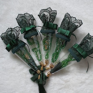May include: Five clear glass tubes filled with green beads and gold accents. Each tube is topped with dark green lace and a matching bow. A dark green tassel hangs from the bottom of the tubes, creating a decorative display.