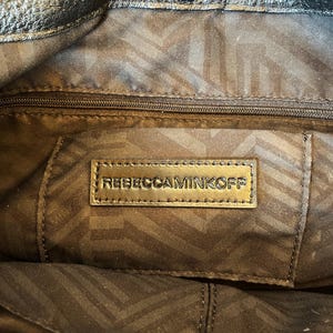 May include: Close-up of the inside of a black leather handbag with a gold zipper and a brown fabric lining. The lining has a geometric pattern. A gold leather tag with the text "REBECCAMINKOFF" is visible.