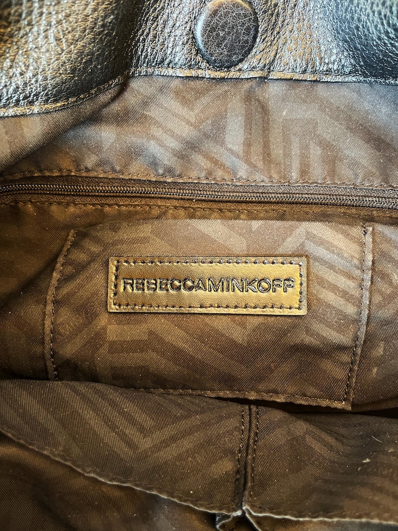 May include: Close-up of the inside of a black leather handbag with a gold zipper and a brown fabric lining. The lining has a geometric pattern. A gold leather tag with the text "REBECCAMINKOFF" is visible.