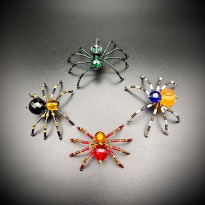 Pode incluir: Quatro ornamentos de aranha de contas com corpos e pernas de diferentes cores. As aranhas são pretas, verdes, vermelhas e azuis.