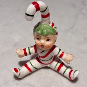 May include: A small ceramic figurine of a seated character wearing a white suit with red and green stripes. The character has a green cap that extends upward into a curved candy cane shape, with painted facial features including eyes and a mouth.