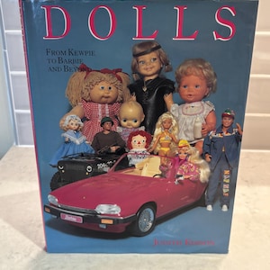 May include: A book cover titled "DOLLS" with the subtitle "From Kewpie to Barbie and Beyond." The cover features a collection of dolls, including a Cabbage Patch doll, and a pink convertible car with a Barbie license plate.