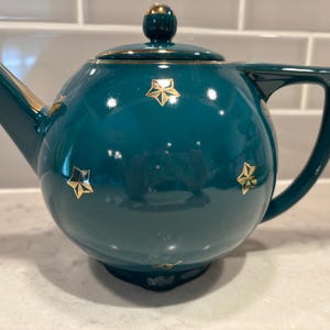 May include: Teal ceramic teapot with a gold star pattern. The teapot has a rounded body, a curved handle, and a spout. The lid and the top of the handle are accented with gold. The teapot is sitting on a white surface.